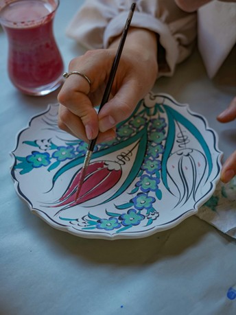 atelier-ceramique-printemps.jpg Main tenant un pinceau décorant une assiette en céramique avec des motifs floraux colorés, dans un atelier de peinture artistique.