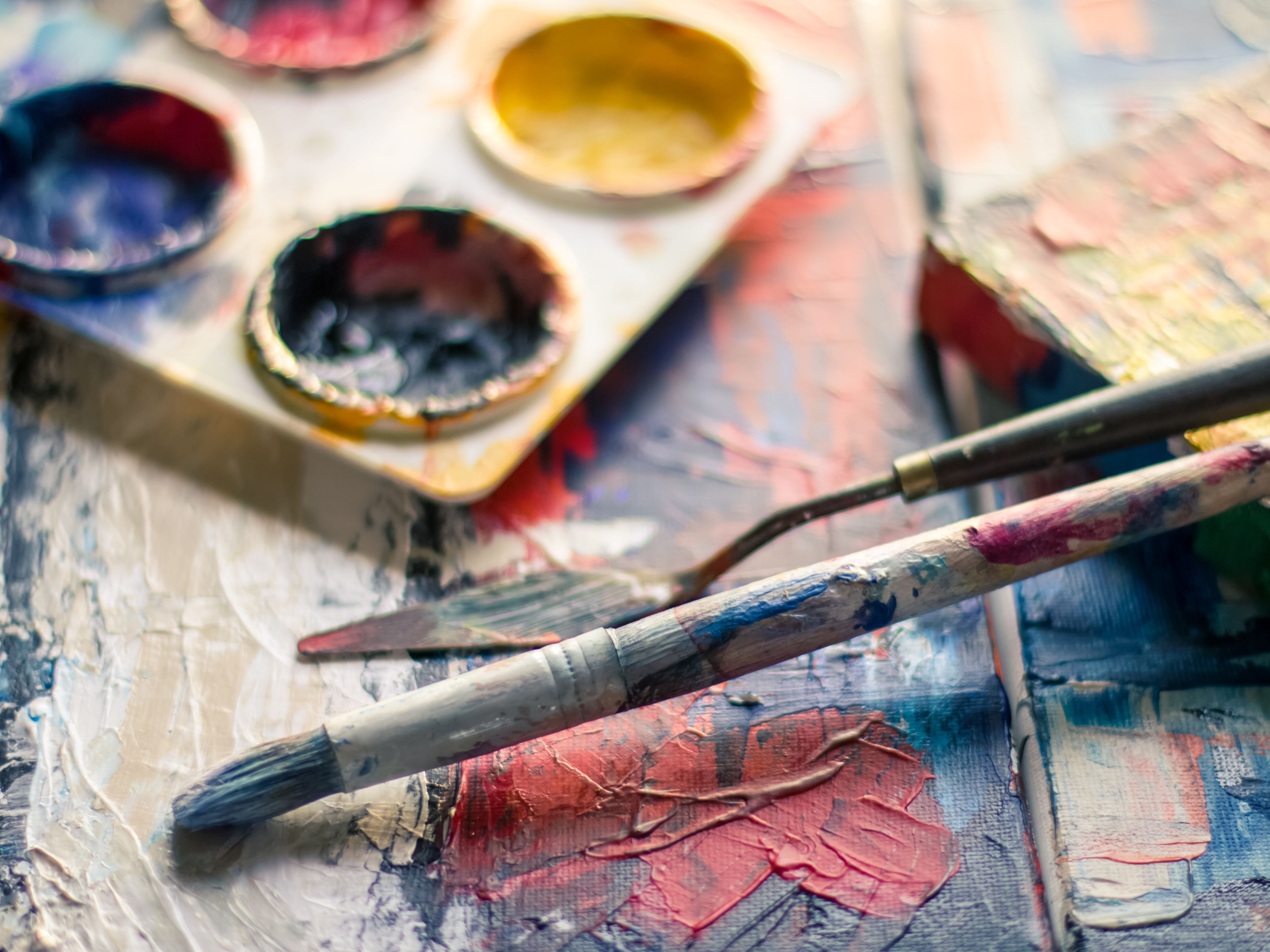 Palette de gouache ouverte avec des godets de couleurs, pinceaux et spatules posés sur une surface peinte aux textures épaisses et multicolores.