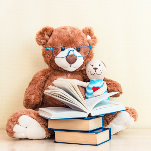 Peluche en forme d’ours assise derrière un livre ouvert, avec une petite peluche à côté et une pile de livres au premier plan.
