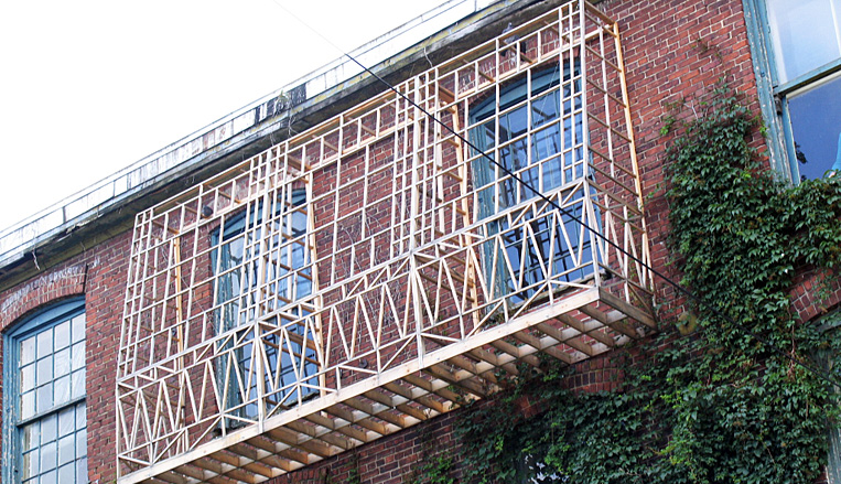 projets-art-infiltrer-habiter-speculer.jpg Structure en bois fixée sur la façade d’un bâtiment en briques, entourée de fenêtres et de végétation grimpante.