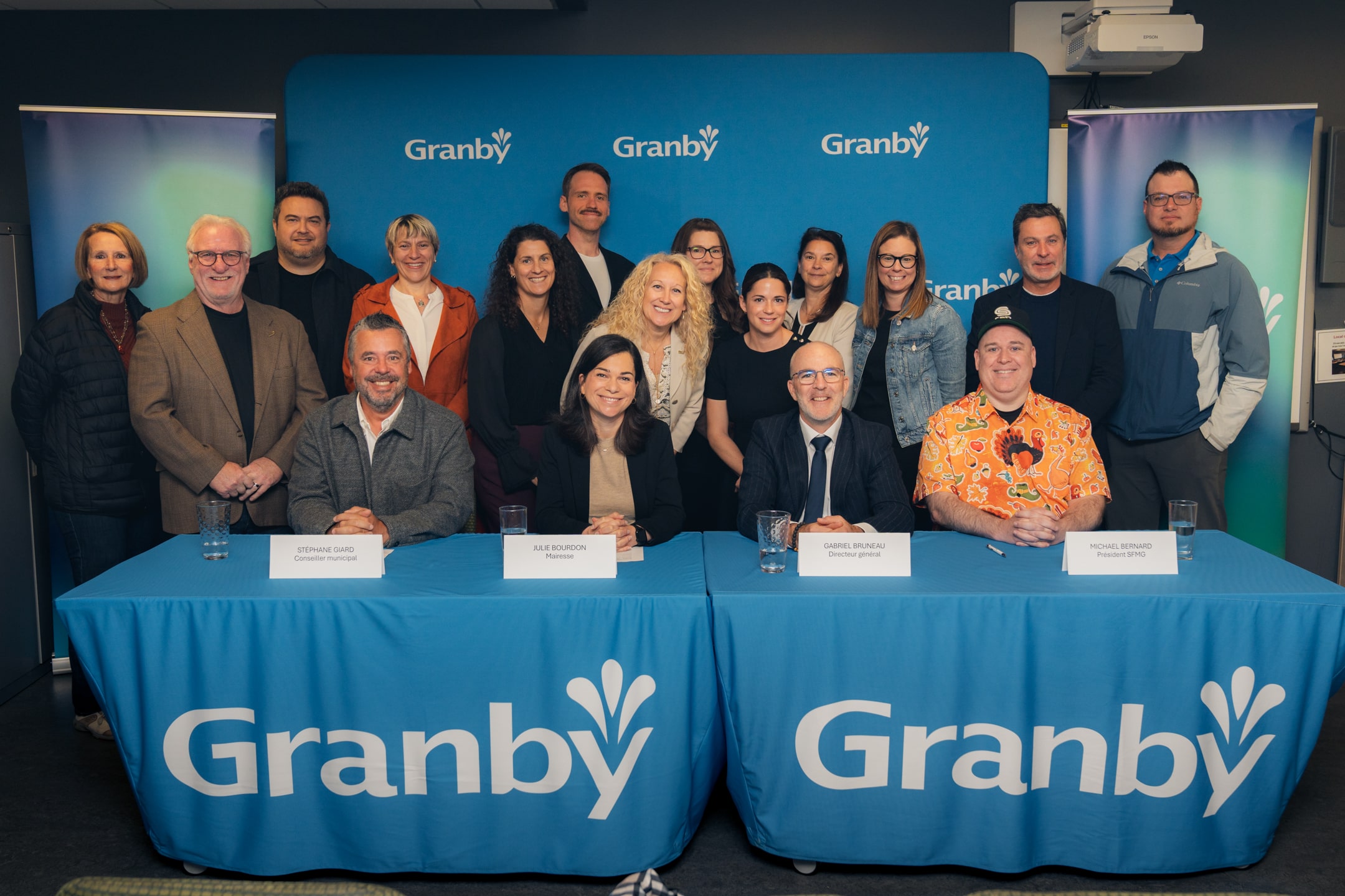 Groupe élargi de personnes posant derrière une table avec nappe bleue “Granby”, trois personnes assises devant des cartons nominatifs.