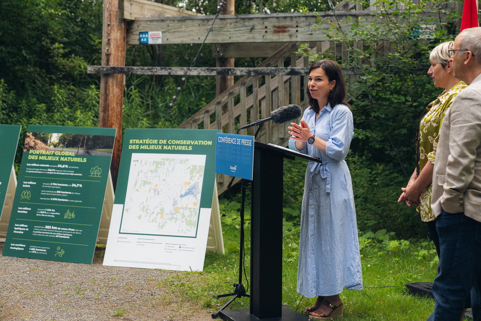 Julie Bourdon mairesse de Granby, derrière un lutrin bleu portant l’inscription “Conférence de presse Granby”, en extérieur, avec des drapeaux du Québec et du Canada en arrière-plan, lors de la présentation de la Stratégie pour un avenir vert et durable.