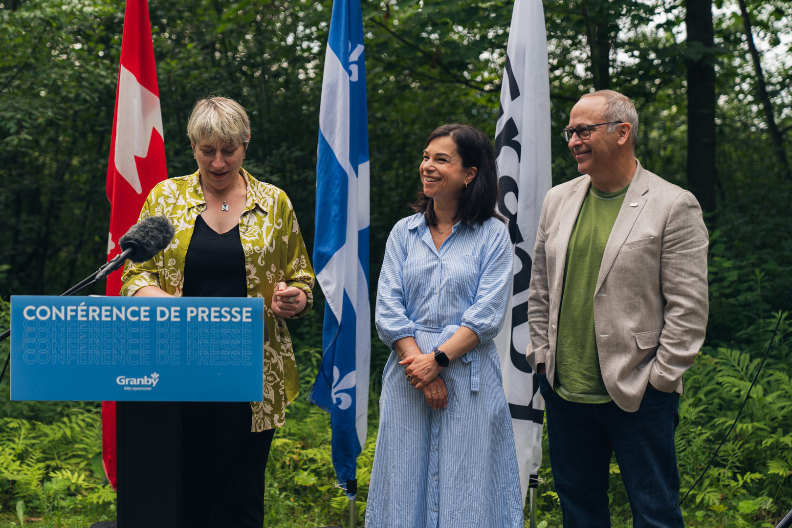 Personne s’exprimant à un micro derrière un lutrin bleu portant l’inscription “Conférence de presse Granby”, en extérieur, avec des drapeaux du Québec et du Canada en arrière-plan, lors de la présentation de la Stratégie pour un avenir vert et durable.