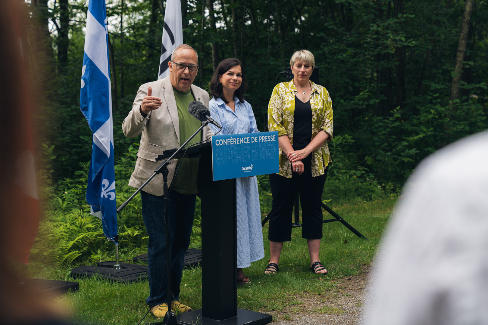 Personne s’exprimant à un micro derrière un lutrin bleu portant l’inscription “Conférence de presse Granby”, en extérieur, avec des drapeaux du Québec et du Canada en arrière-plan, lors de la présentation de la Stratégie pour un avenir vert et durable.
