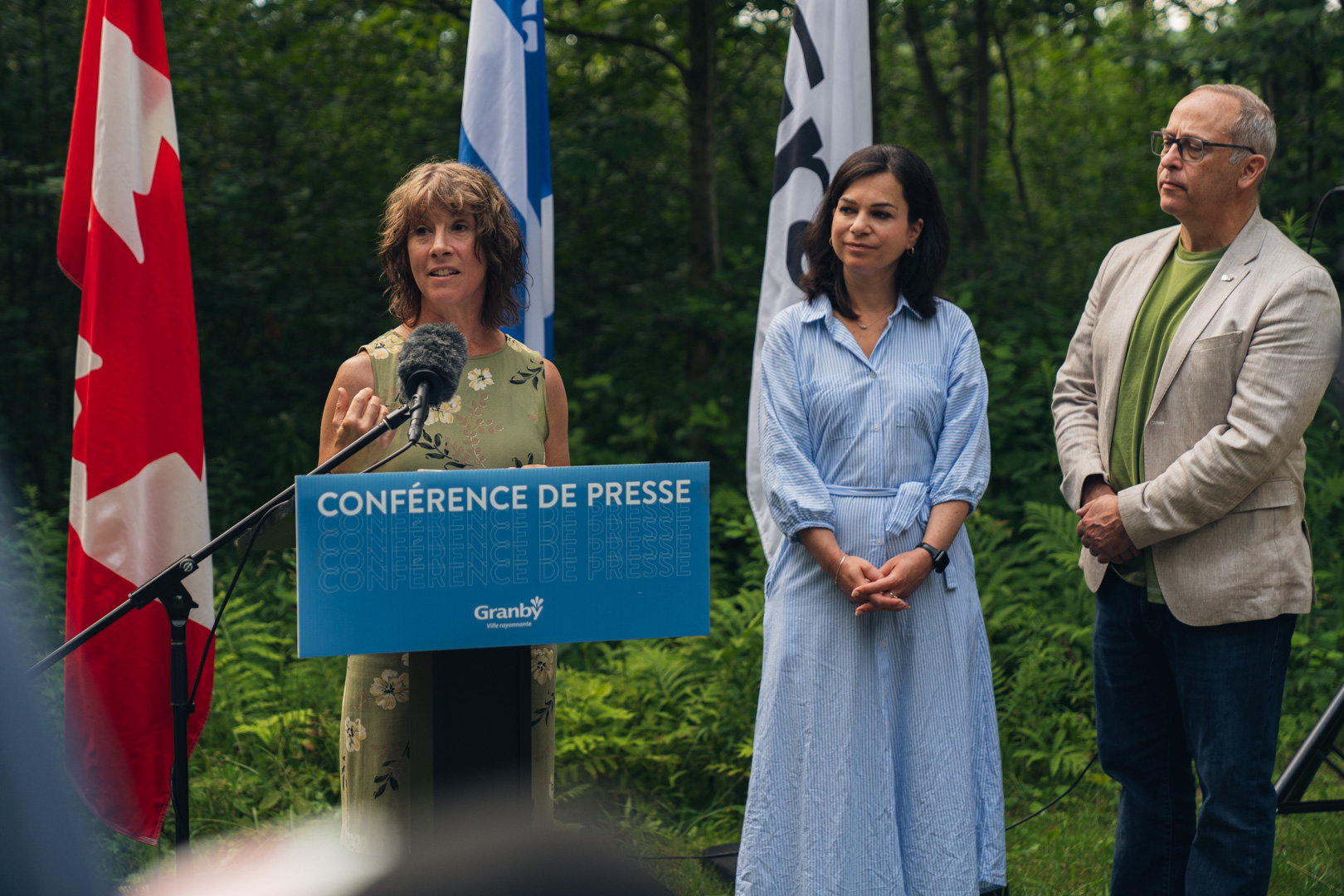 Personne s’exprimant à un micro derrière un lutrin bleu portant l’inscription “Conférence de presse Granby”, en extérieur, avec des drapeaux du Québec et du Canada en arrière-plan, lors de la présentation de la Stratégie pour un avenir vert et durable.