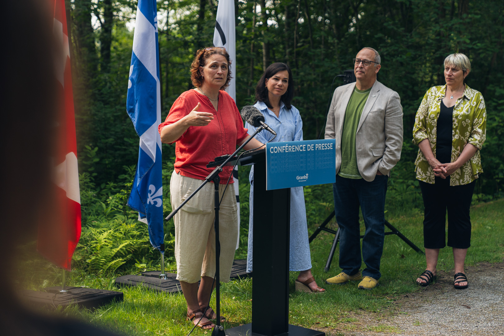 Personne s’exprimant à un micro derrière un lutrin bleu portant l’inscription “Conférence de presse Granby”, en extérieur, avec des drapeaux du Québec et du Canada en arrière-plan, lors de la présentation de la Stratégie pour un avenir vert et durable.