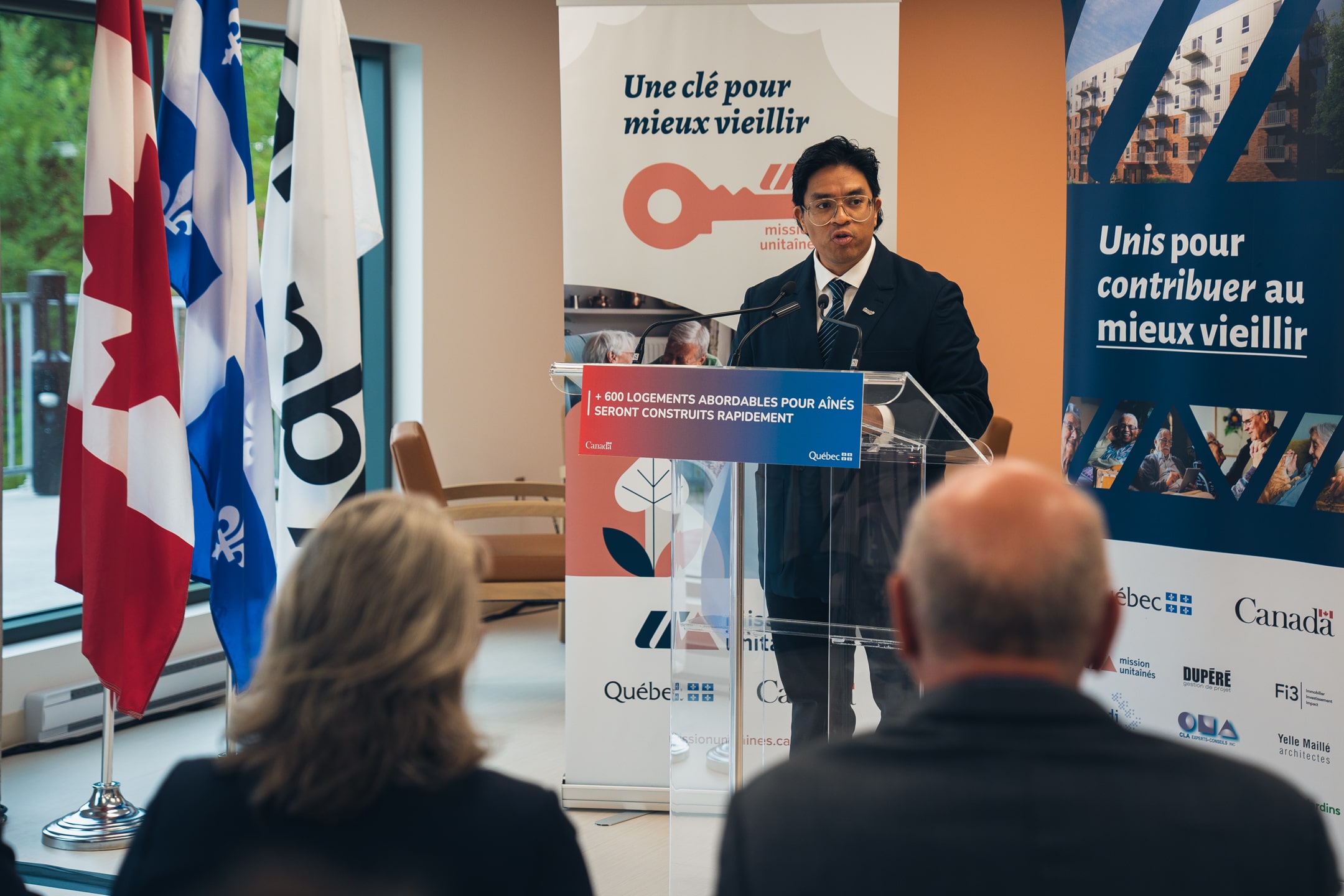 Personne s’exprimant à un micro derrière un lutrin transparent portant l’inscription “600 logements abordables pour aînés seront construits rapidement”, avec des drapeaux du Canada et du Québec en arrière-plan et une bannière Mission Unitaînés affichant le slogan “Une clé pour mieux vieillir”.