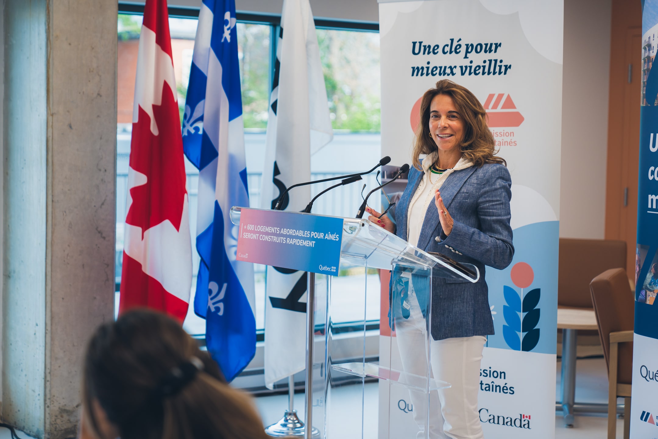 Personne s’exprimant à un micro derrière un lutrin transparent portant l’inscription “600 logements abordables pour aînés seront construits rapidement”, avec des drapeaux du Canada et du Québec en arrière-plan et une bannière Mission Unitaînés affichant le slogan “Une clé pour mieux vieillir”.
