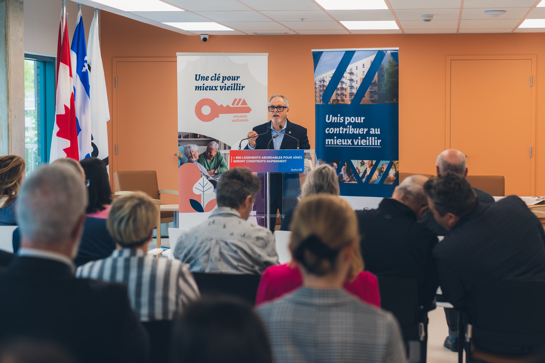 Personne s’exprimant à un micro derrière un lutrin transparent portant l’inscription “600 logements abordables pour aînés seront construits rapidement”, avec des drapeaux du Canada et du Québec en arrière-plan et une bannière Mission Unitaînés affichant le slogan “Une clé pour mieux vieillir”.