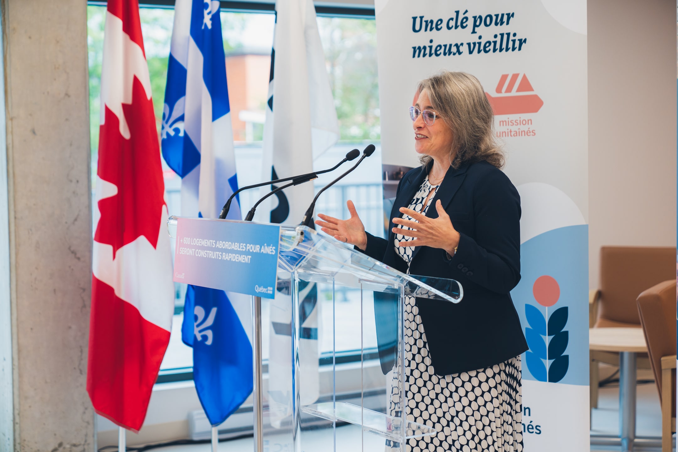 Personne s’exprimant à un micro derrière un lutrin transparent portant l’inscription “600 logements abordables pour aînés seront construits rapidement”, avec des drapeaux du Canada et du Québec en arrière-plan et une bannière Mission Unitaînés affichant le slogan “Une clé pour mieux vieillir”.