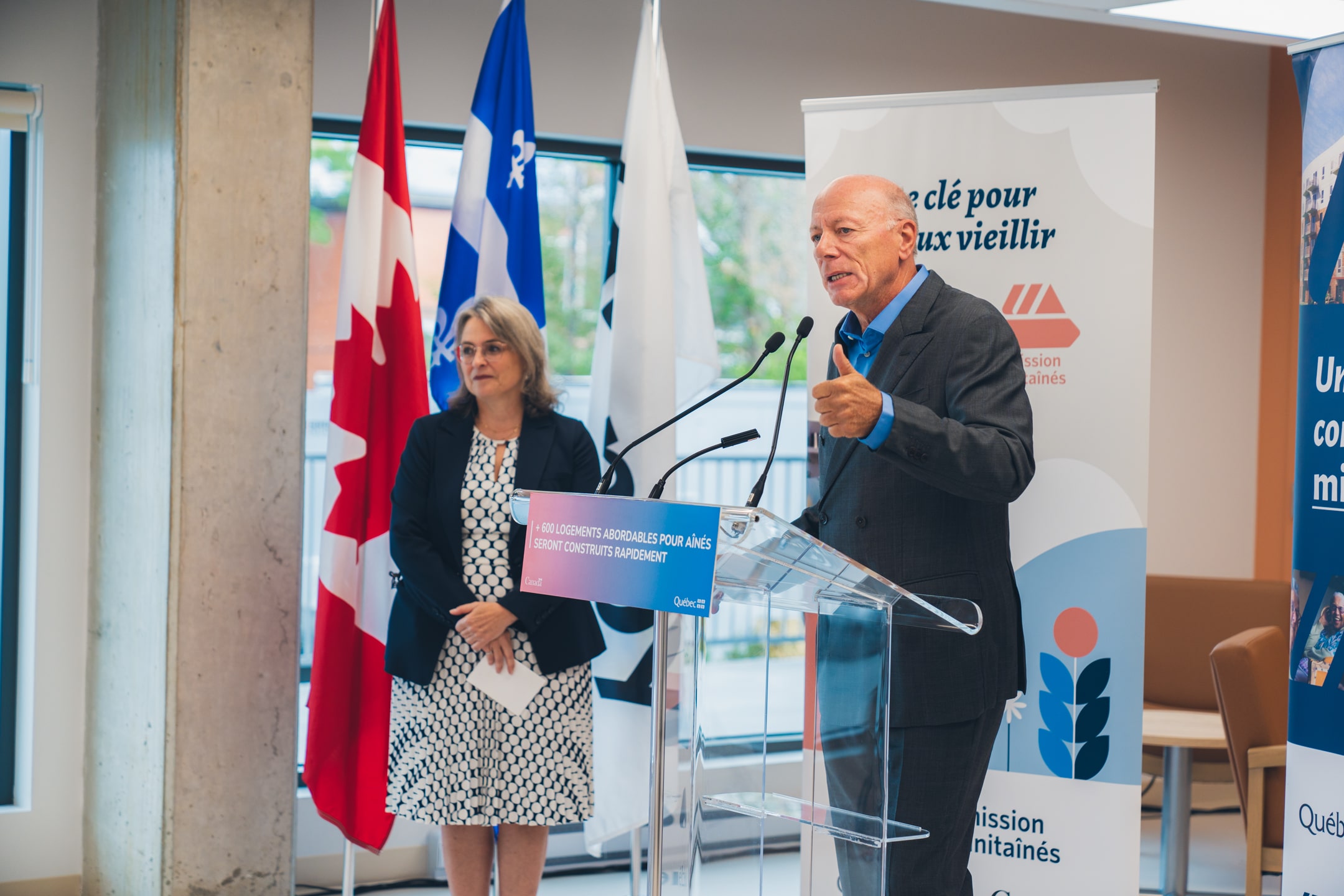 Personne s’exprimant à un micro derrière un lutrin transparent portant l’inscription “600 logements abordables pour aînés seront construits rapidement”, avec des drapeaux du Canada et du Québec en arrière-plan et une bannière Mission Unitaînés affichant le slogan “Une clé pour mieux vieillir”.