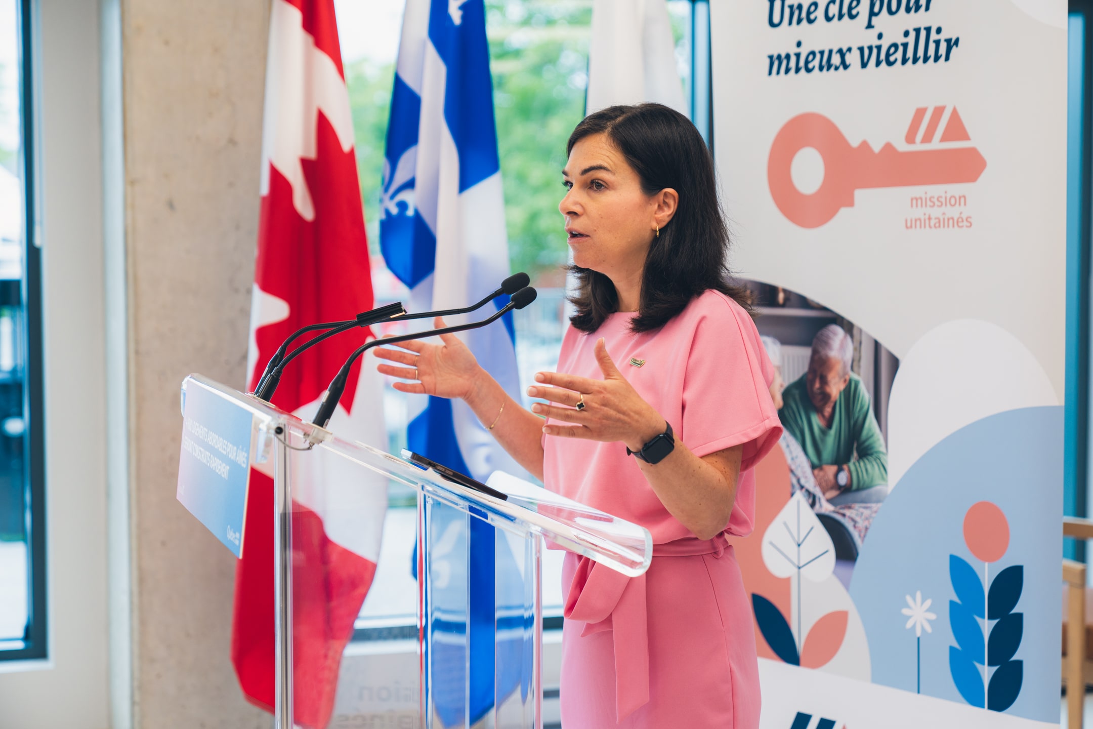 Julie Bourdon mairesse de Granby s’exprimant à un micro derrière un lutrin transparent portant l’inscription “600 logements abordables pour aînés seront construits rapidement”, avec des drapeaux du Canada et du Québec en arrière-plan et une bannière Mission Unitaînés affichant le slogan “Une clé pour mieux vieillir”.