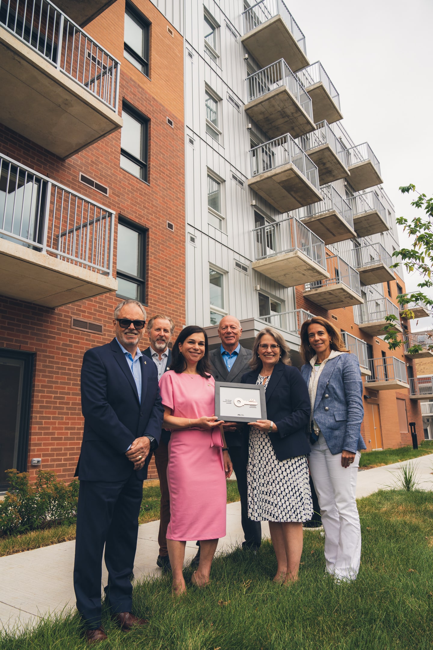 Julie Bourdon mairesse de Granby  etr un groupe de personnes debout devant un immeuble résidentiel moderne en brique et aluminium, avec plusieurs balcons, tenant une plaque symbolisant la remise des clés lors d’un événement officiel. 