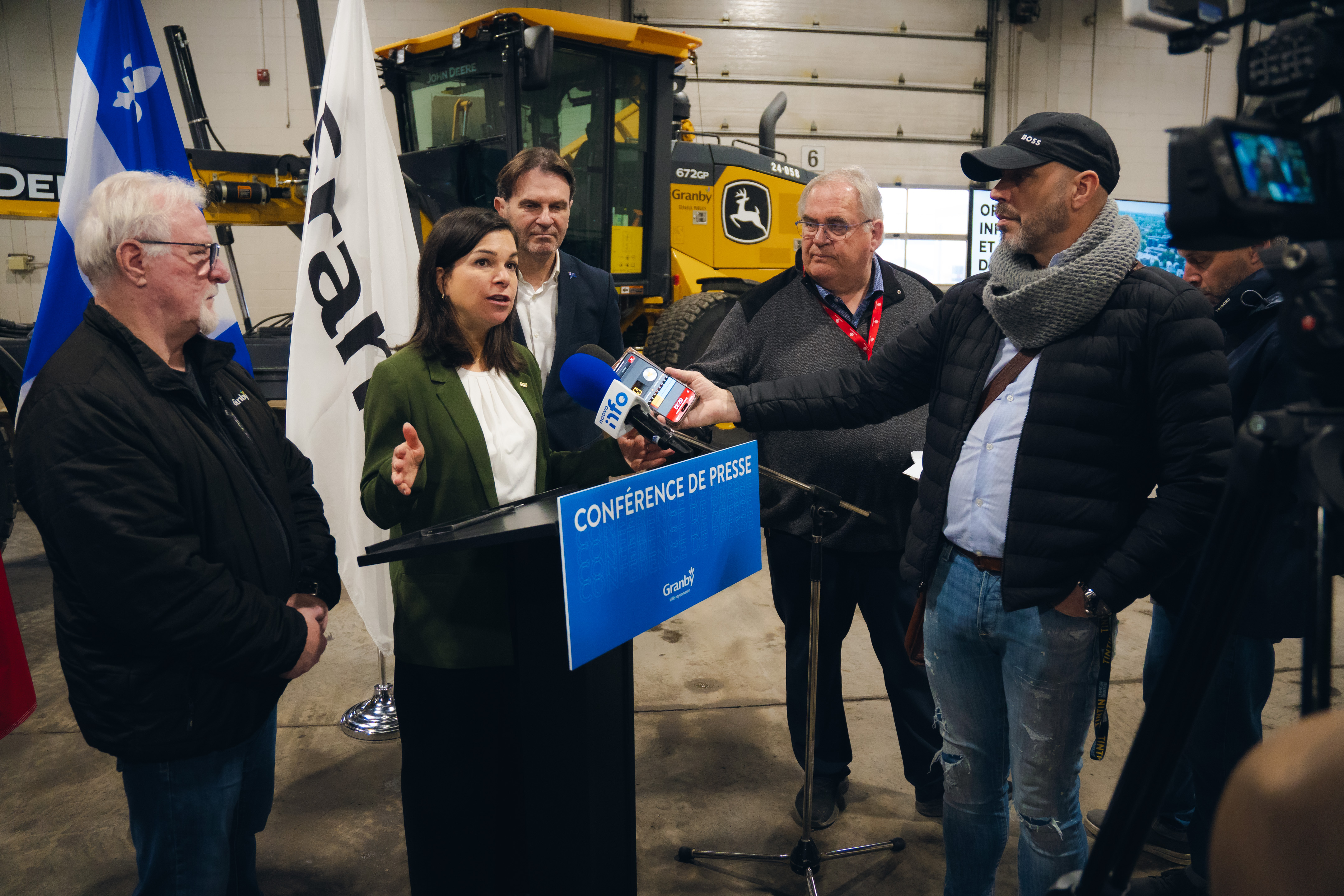 Conférence de presse de Julie Bourdon mairesse de Granby dans un garage municipal avec un podium, des micros de médias, des drapeaux et un véhicule de voirie en arrière-plan.