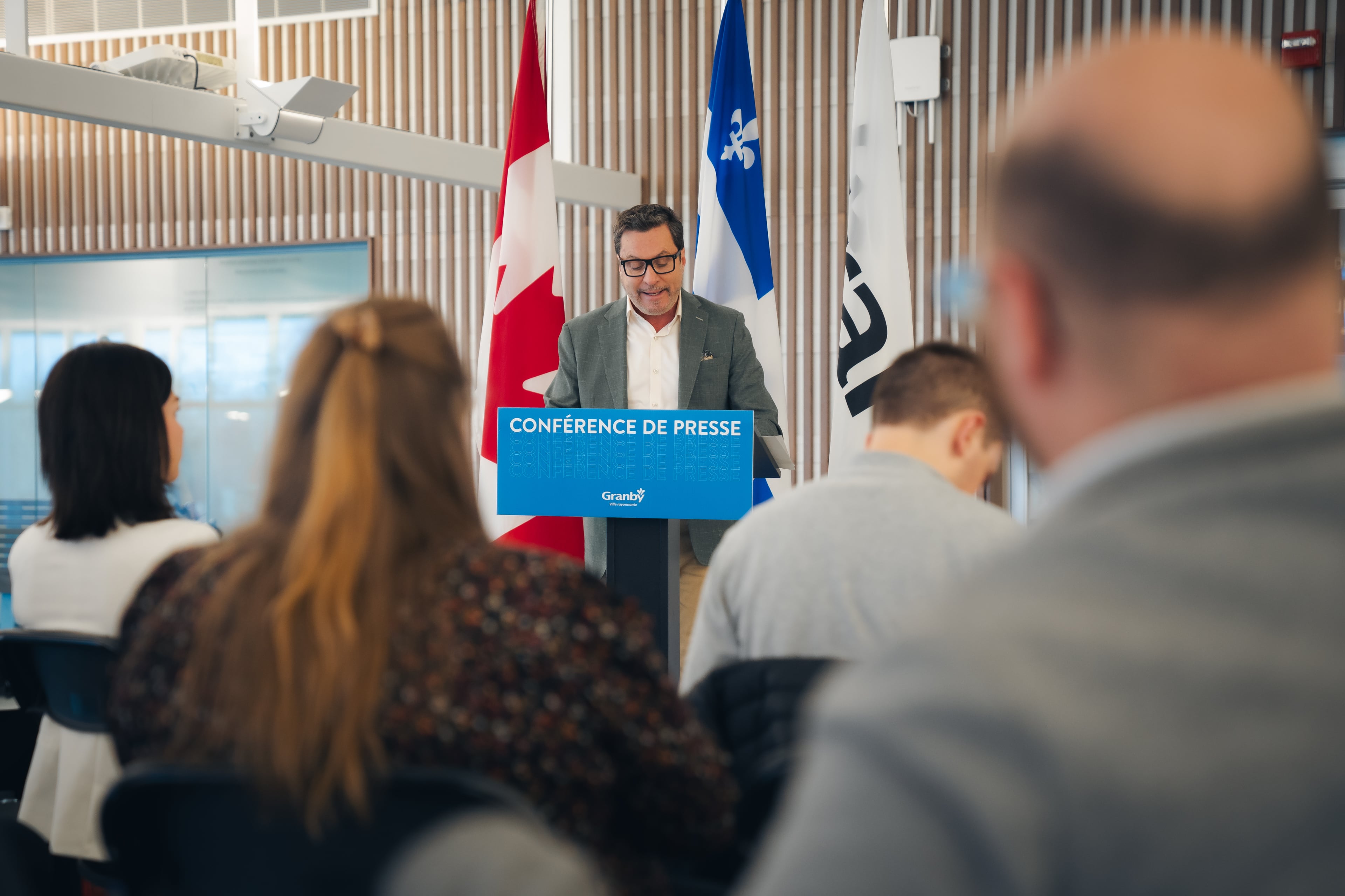 Personne debout derrière un lutrin bleu portant l’inscription “Conférence de presse” à l’intérieur d’une salle moderne. Quelques personnes sont assises en avant-plan, écoutant la présentation.