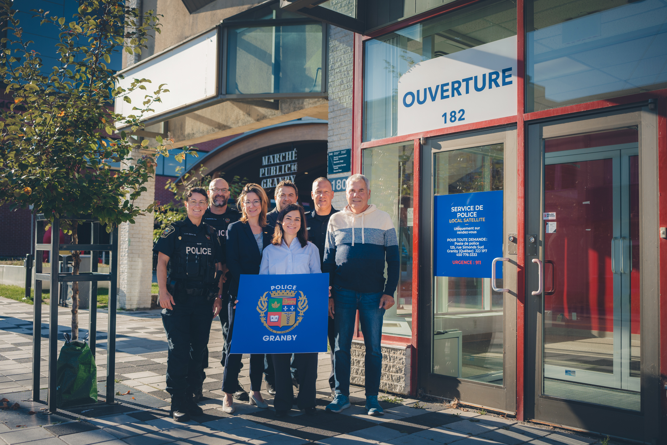 Julie Bourdon mairesse de Granby, avec un groupe de personnes debout devant la façade vitrée d’un nouveau local de police satellite au centre-ville, avec une affiche bleue indiquant “Police Granby” et une porte portant la mention Ouverture 182.
