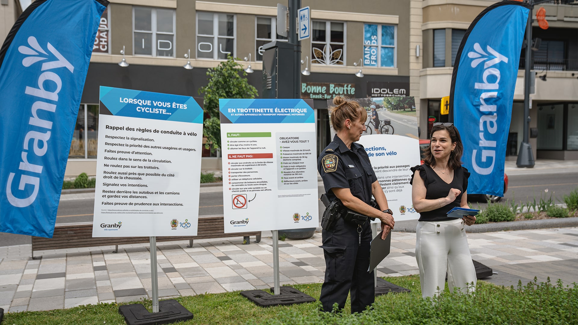 Julie Bourdon mairesse de Granby, dont un agents de police en uniforme, posent devant des panneaux éducatifs sur la sécurité des cyclistes et des trottinettes dans un espace urbain à Granby.
