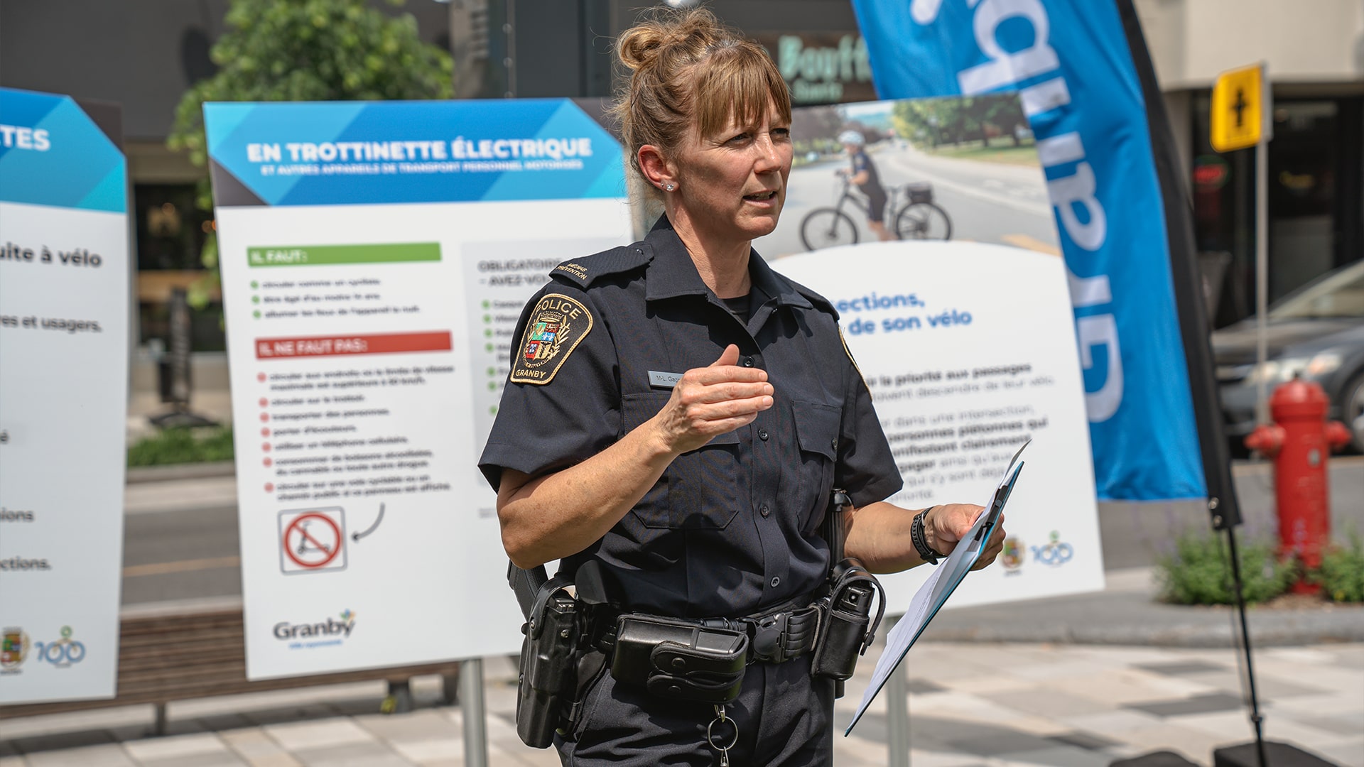 Une agents de police en uniforme, posent devant des panneaux éducatifs sur la sécurité des cyclistes et des trottinettes dans un espace urbain à Granby. 