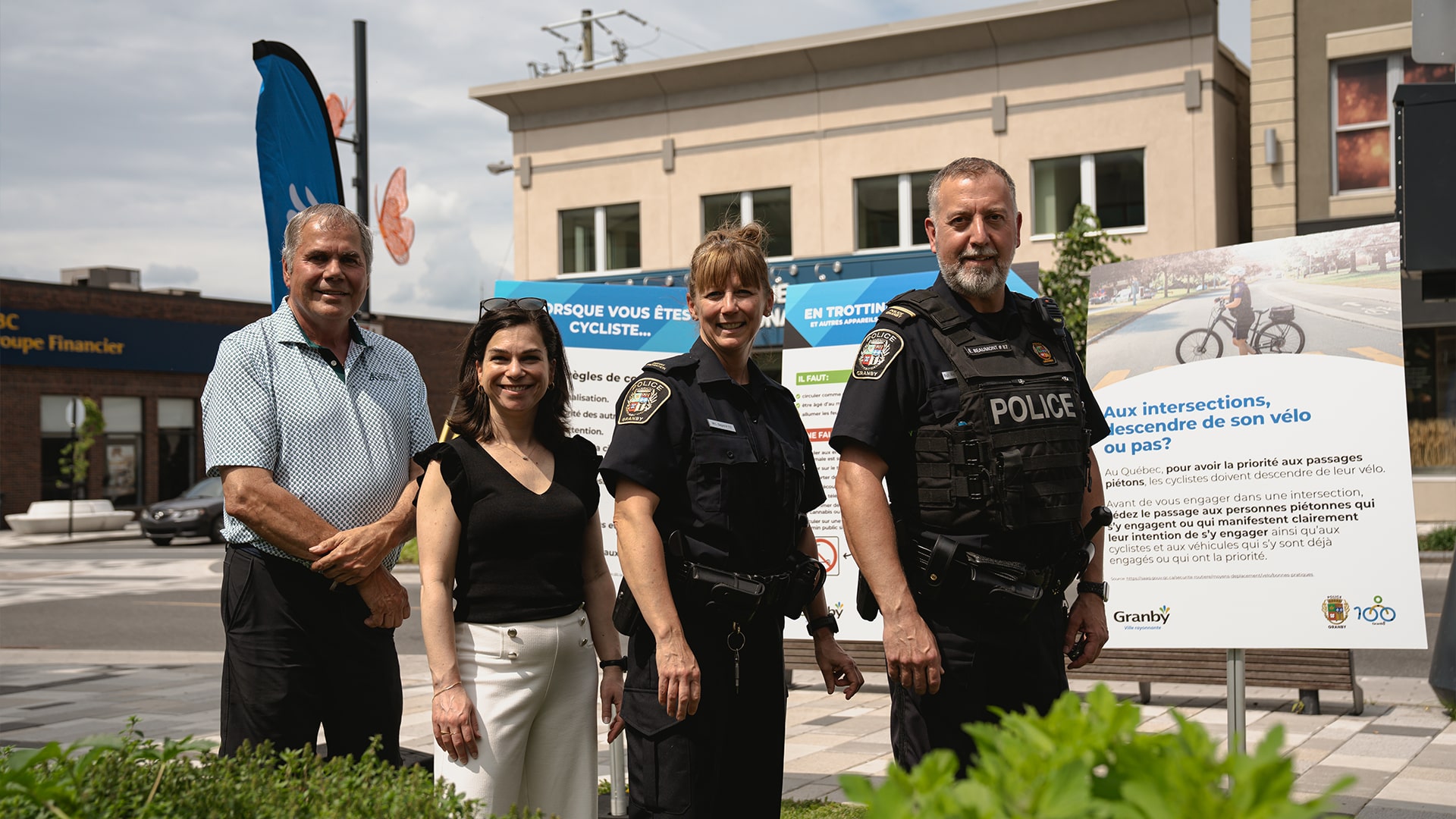 Julie Bourdon mairesse de Granby et quatre personnes, dont deux agents de police en uniforme, posent devant des panneaux éducatifs sur la sécurité des cyclistes et des trottinettes dans un espace urbain à Granby. 