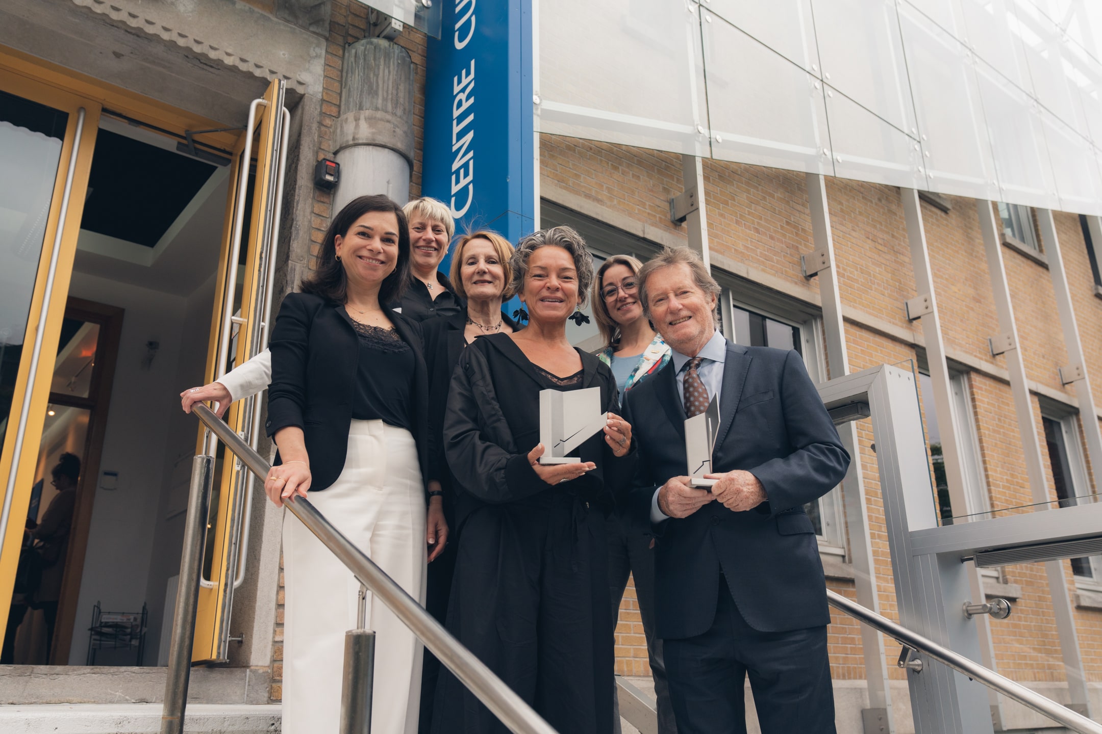 Groupe de personnes posant sur les marches à l’extérieur d’un bâtiment avec trophées en main, devant une bannière bleue indiquant Centre culturel.