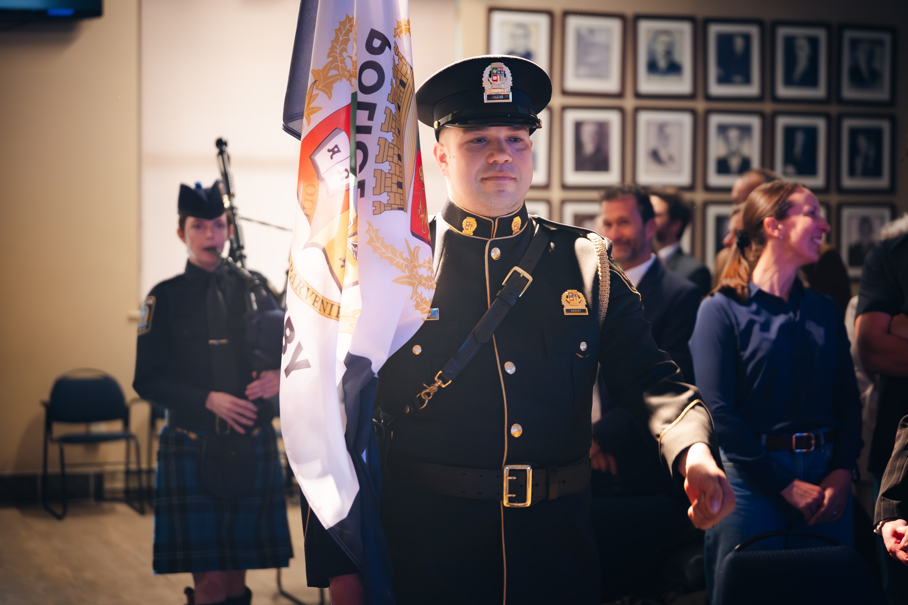 Agent en uniforme cérémonial tenant le drapeau du Service de police de Granby lors d’une cérémonie, avec un joueur de cornemuse en arrière-plan et des personnes assises dans la salle.